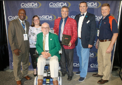 Chris Wenzler (red jacket) posing with fellow winners of the CoSIDA Bob Kenworthy Community Service Award. Bob Kenworthy is seated in the green jacket.